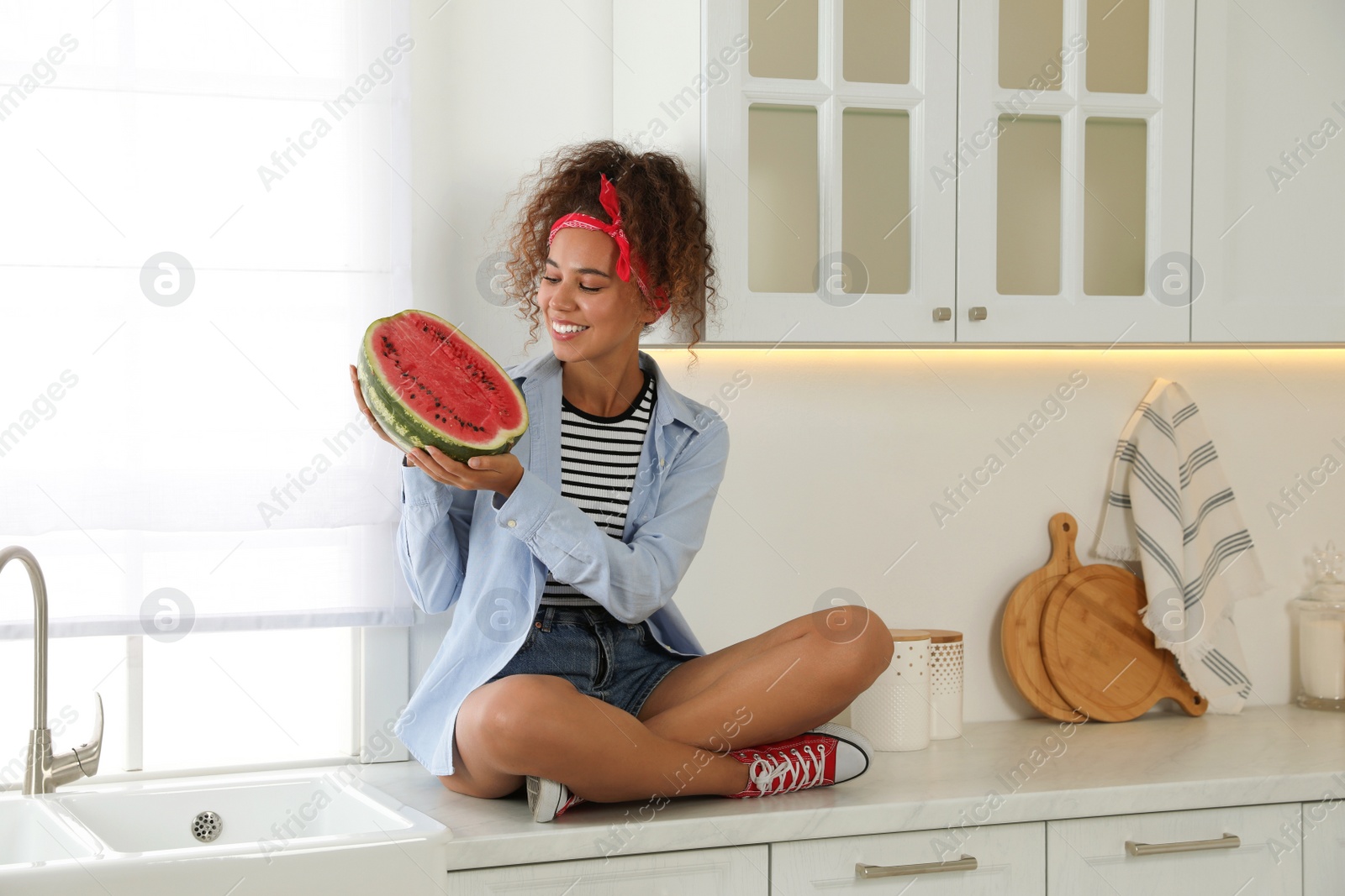 Photo of Beautiful young African American woman with half of watermelon sitting on countertop in kitchen