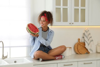 Photo of Beautiful young African American woman with half of watermelon sitting on countertop in kitchen