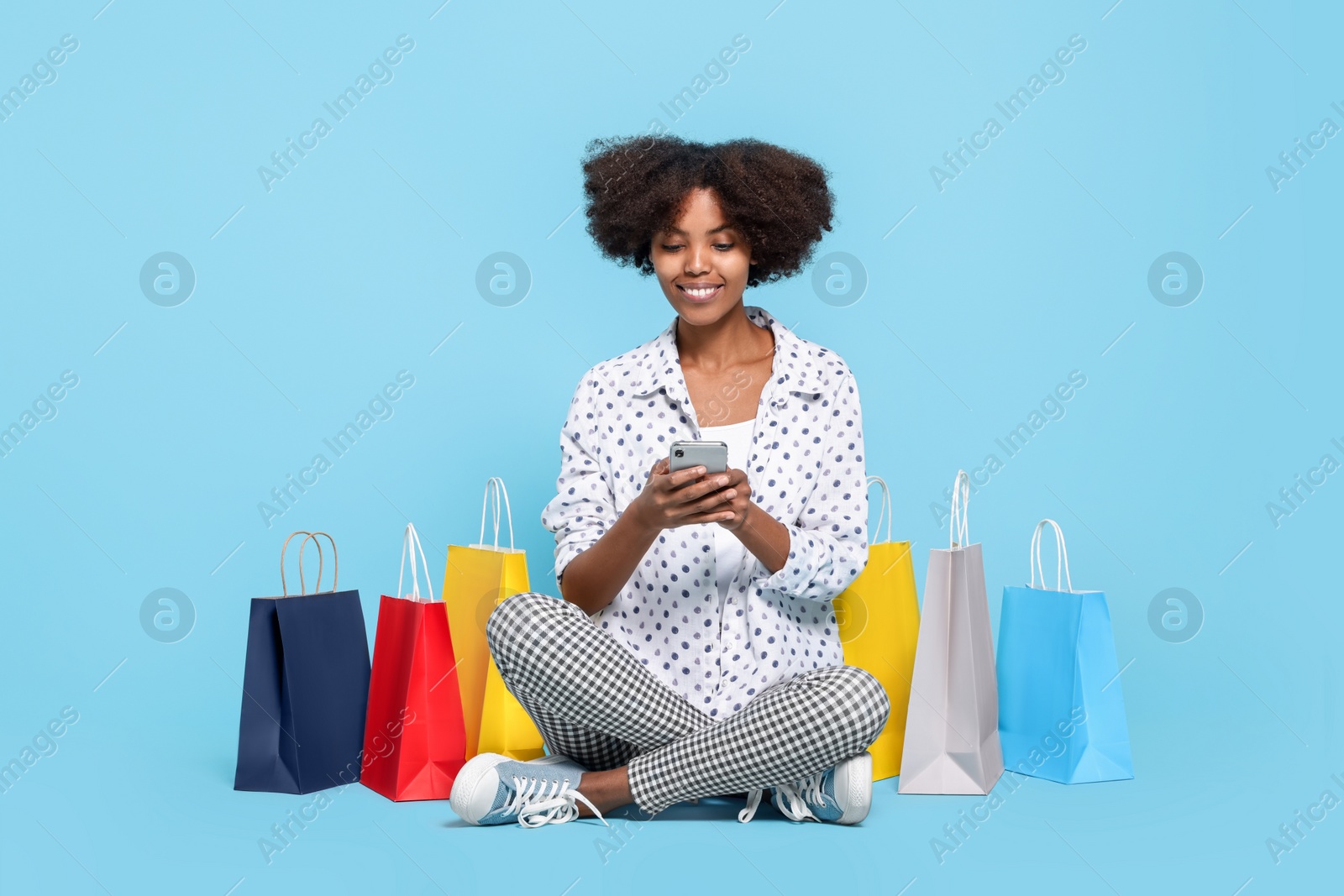 Photo of Happy African American woman with shopping bags and smartphone on light blue background