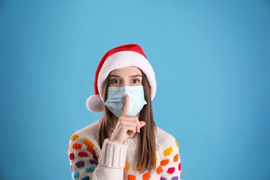 Photo of Pretty woman in Santa hat and medical mask showing silence gesture on light blue background