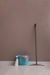 Photo of Floor mop, cleaning detergent and bucket with rag near brown wall indoors
