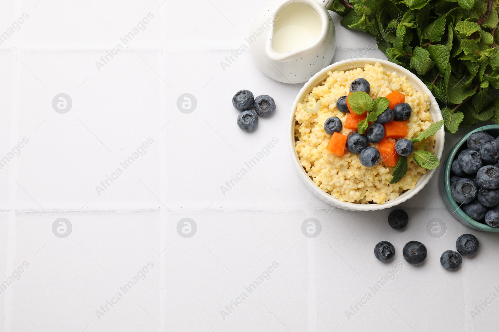 Photo of Tasty millet porridge with blueberries, pumpkin and mint in bowl on white tiled table, flat lay. Space for text