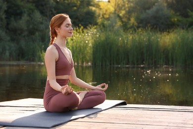 Beautiful woman practicing Padmasana on yoga mat on wooden pier near pond, space for text. Lotus pose
