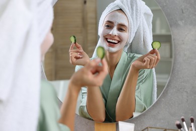 Photo of Woman with face mask and cucumber slices near mirror in bathroom. Spa treatments