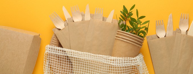 Paper and wooden tableware with green twigs on yellow background, flat lay. Banner design