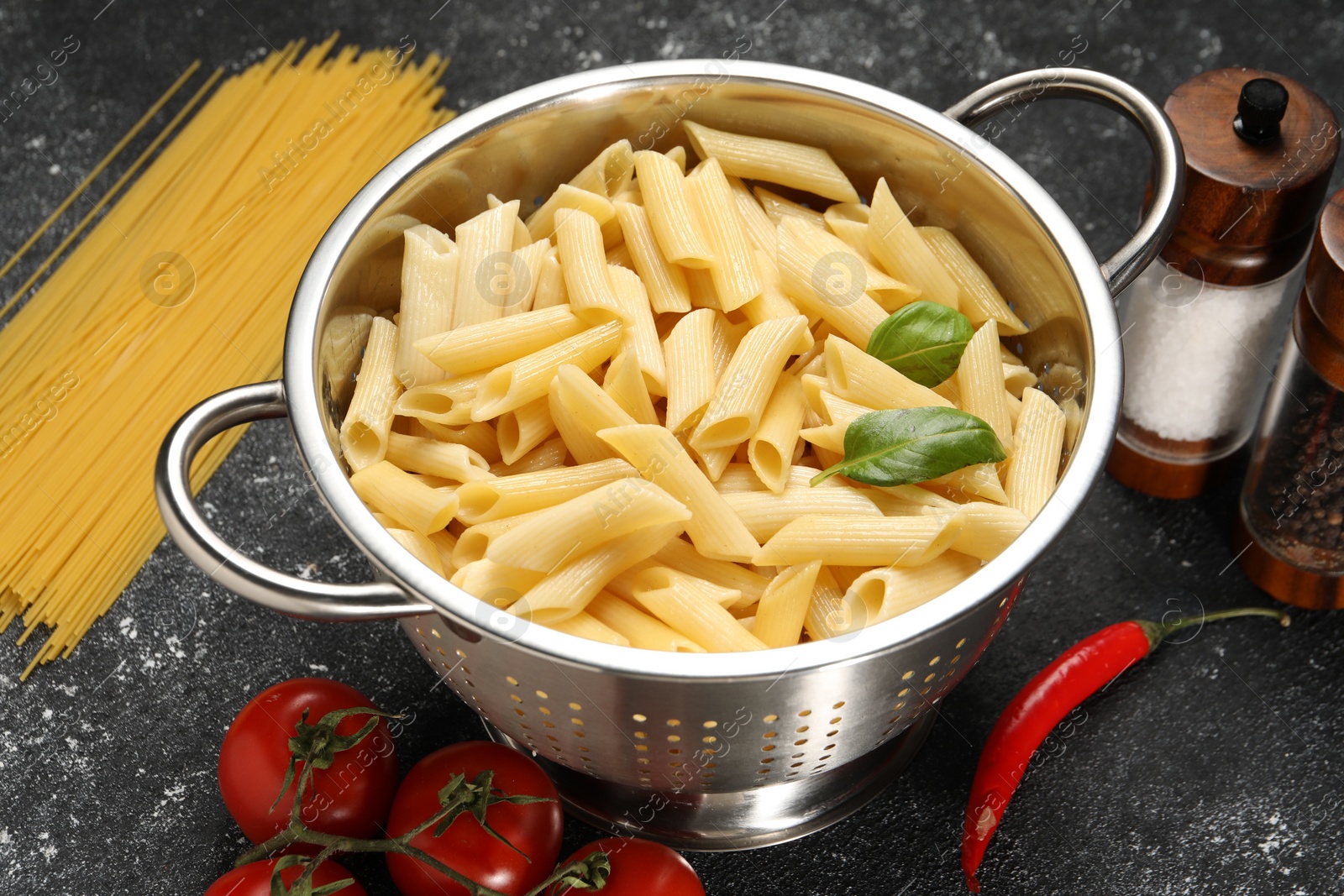 Photo of Cooked pasta in metal colander, products and spices on dark textured table, closeup