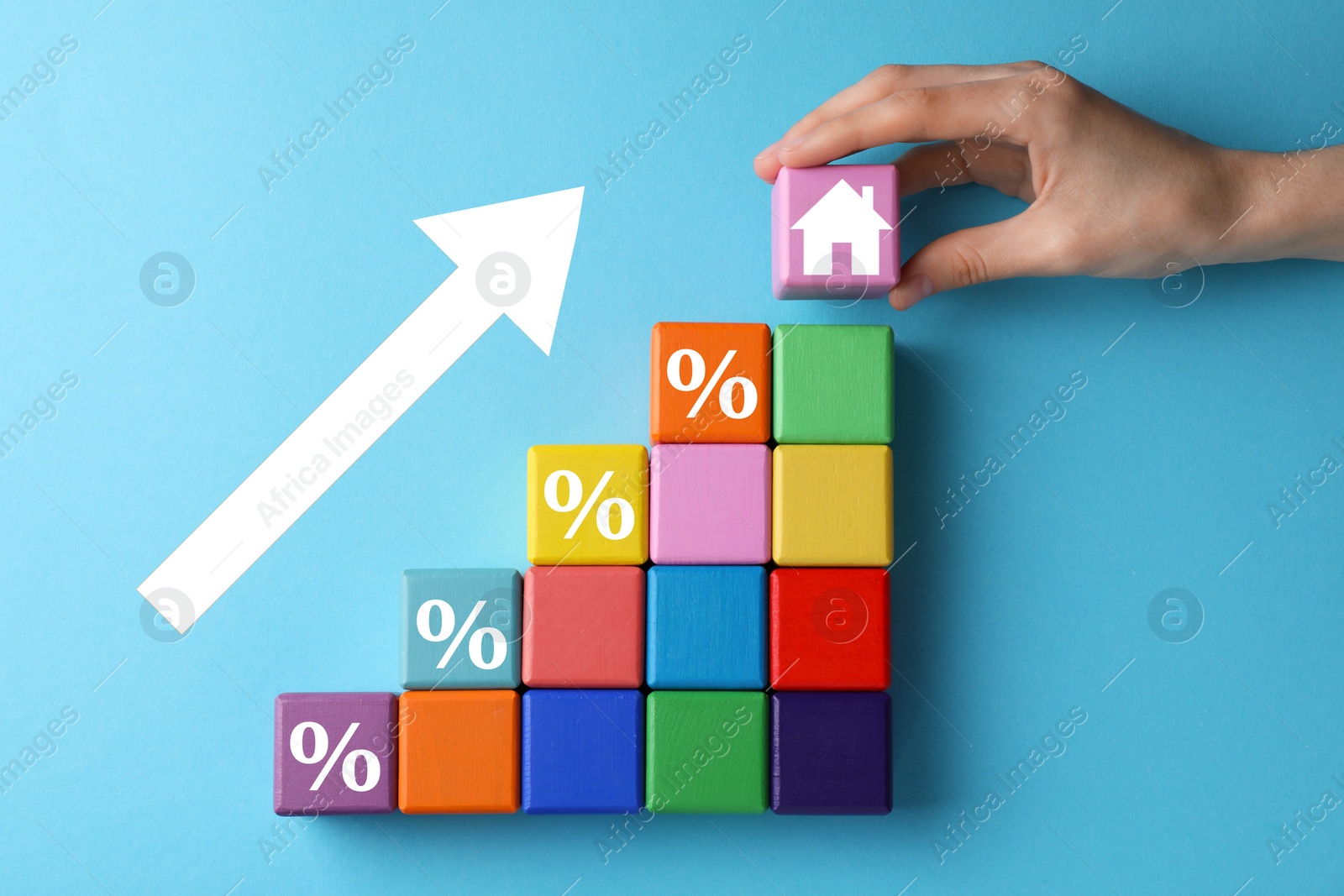 Image of Mortgage rate rising illustrated by upward arrow. Woman putting cube with house icon near other ones on light blue background, top view