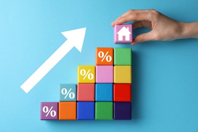 Mortgage rate rising illustrated by upward arrow. Woman putting cube with house icon near other ones on light blue background, top view