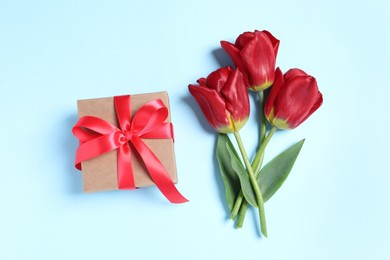 Photo of Beautiful gift box with bow and red tulips on light blue background, flat lay