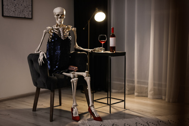 Photo of Skeleton in dress with wine sitting at table indoors