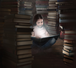 Image of Cute little girl reading on wooden floor in room full of stacked books