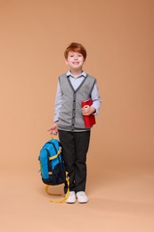 Happy schoolboy with book and backpack on beige background