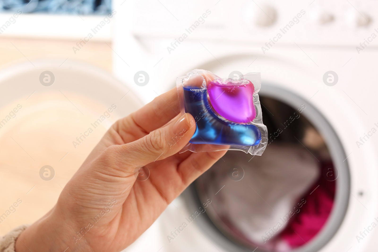 Photo of Woman holding laundry detergent capsule near washing machine indoors, closeup