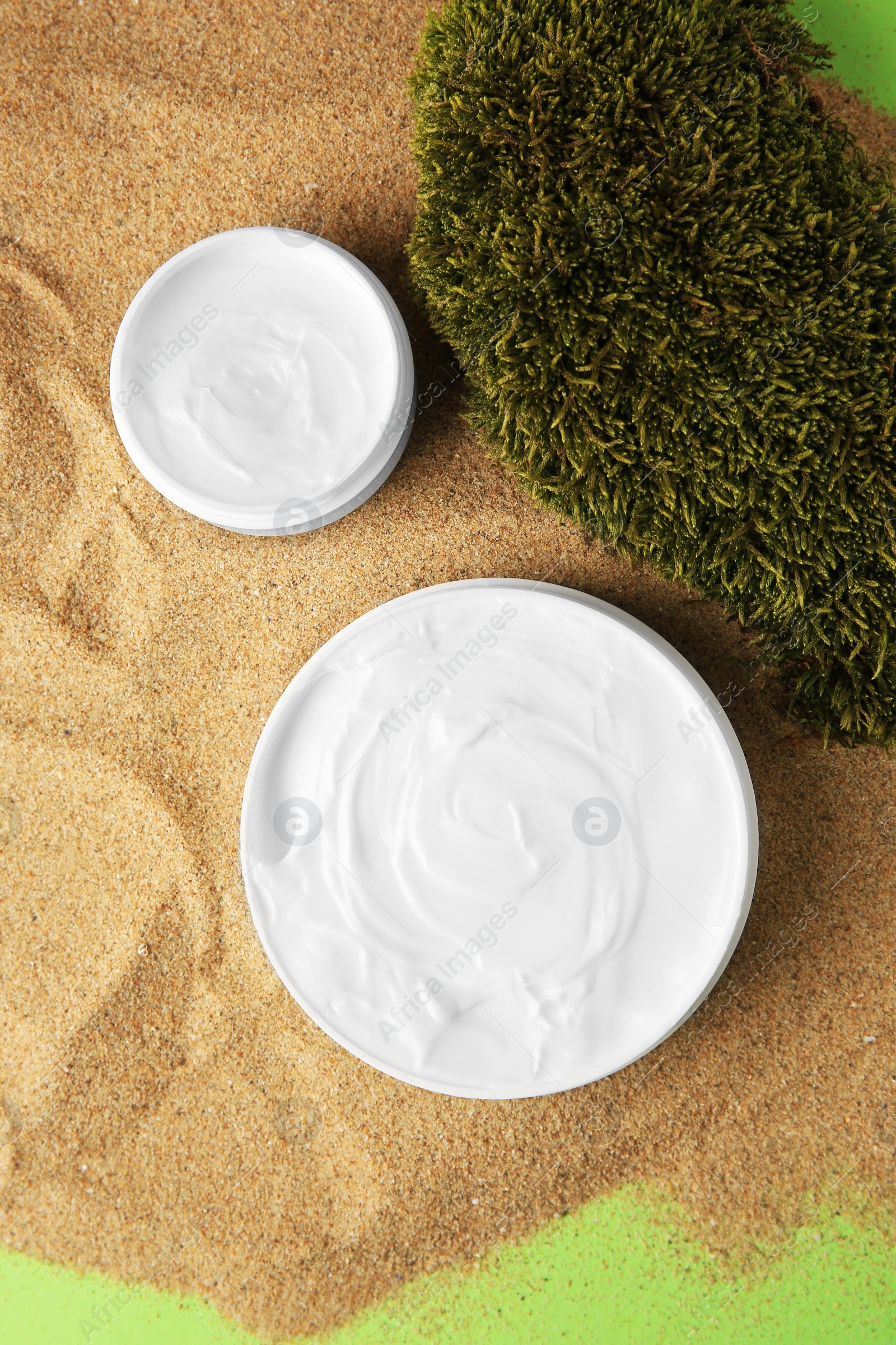 Photo of Jars with cream and moss on sand against green background, flat lay. Cosmetic product