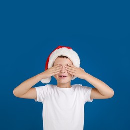 Happy little child in Santa hat closing eyes on blue background. Christmas celebration
