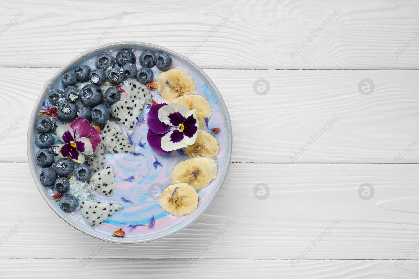 Photo of Delicious smoothie bowl with fresh fruits, blueberries and flowers on white wooden table, top view. Space for text