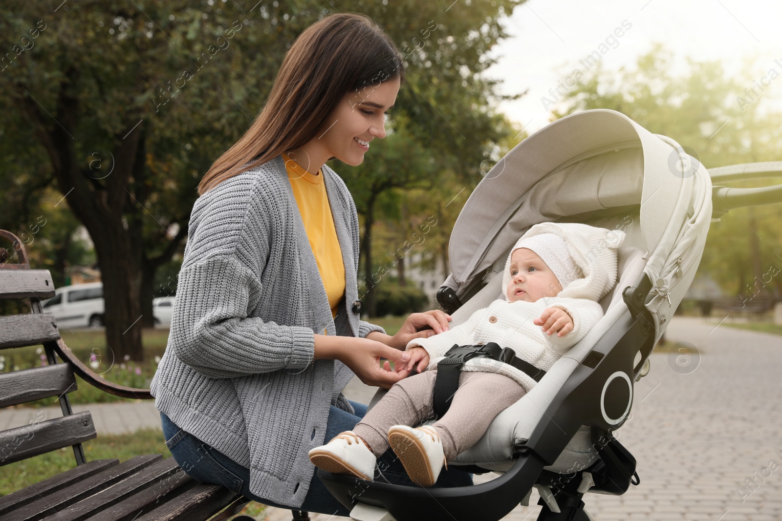 Photo of Happy mother with her adorable baby in stroller sitting on bench outdoors