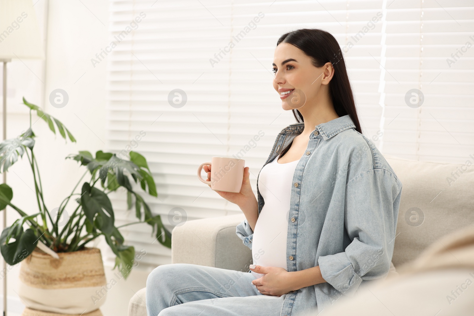 Photo of Happy pregnant woman with cup of drink on sofa at home