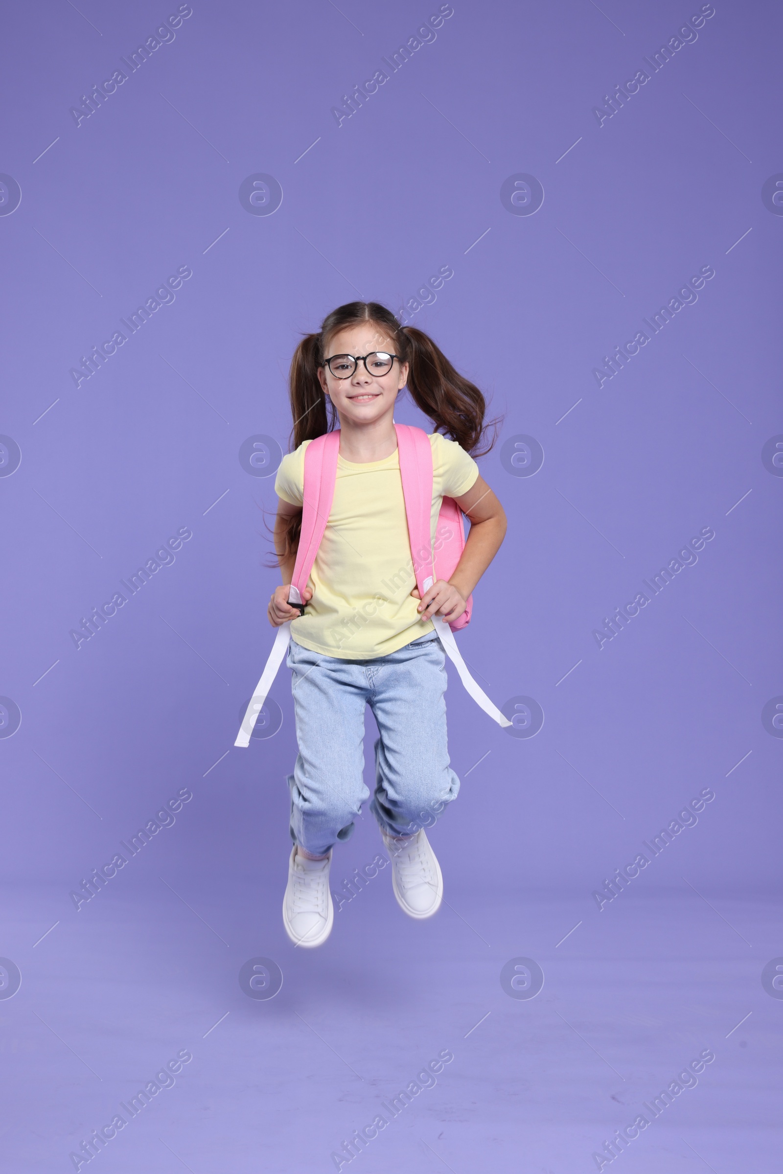 Photo of Cute schoolgirl in glasses with backpack jumping on violet background