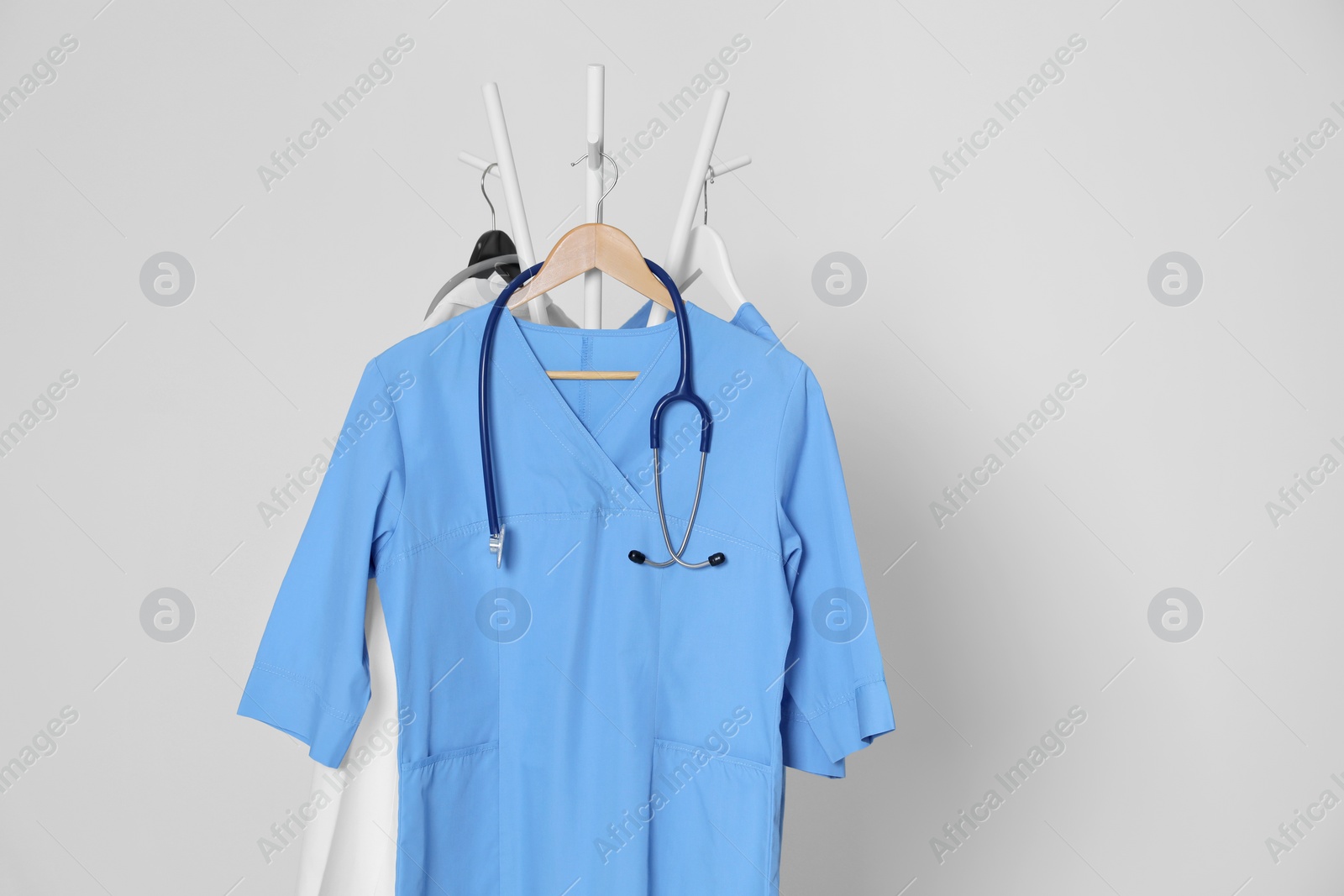 Photo of Medical uniforms and stethoscope hanging on rack against light grey background. Space for text