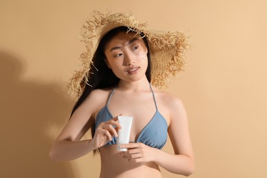 Photo of Beautiful young woman in straw hat with sunscreen on her face holding sun protection cream against beige background