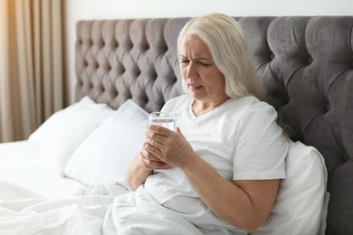 Photo of Mature woman with terrible headache holding glass of water in bed