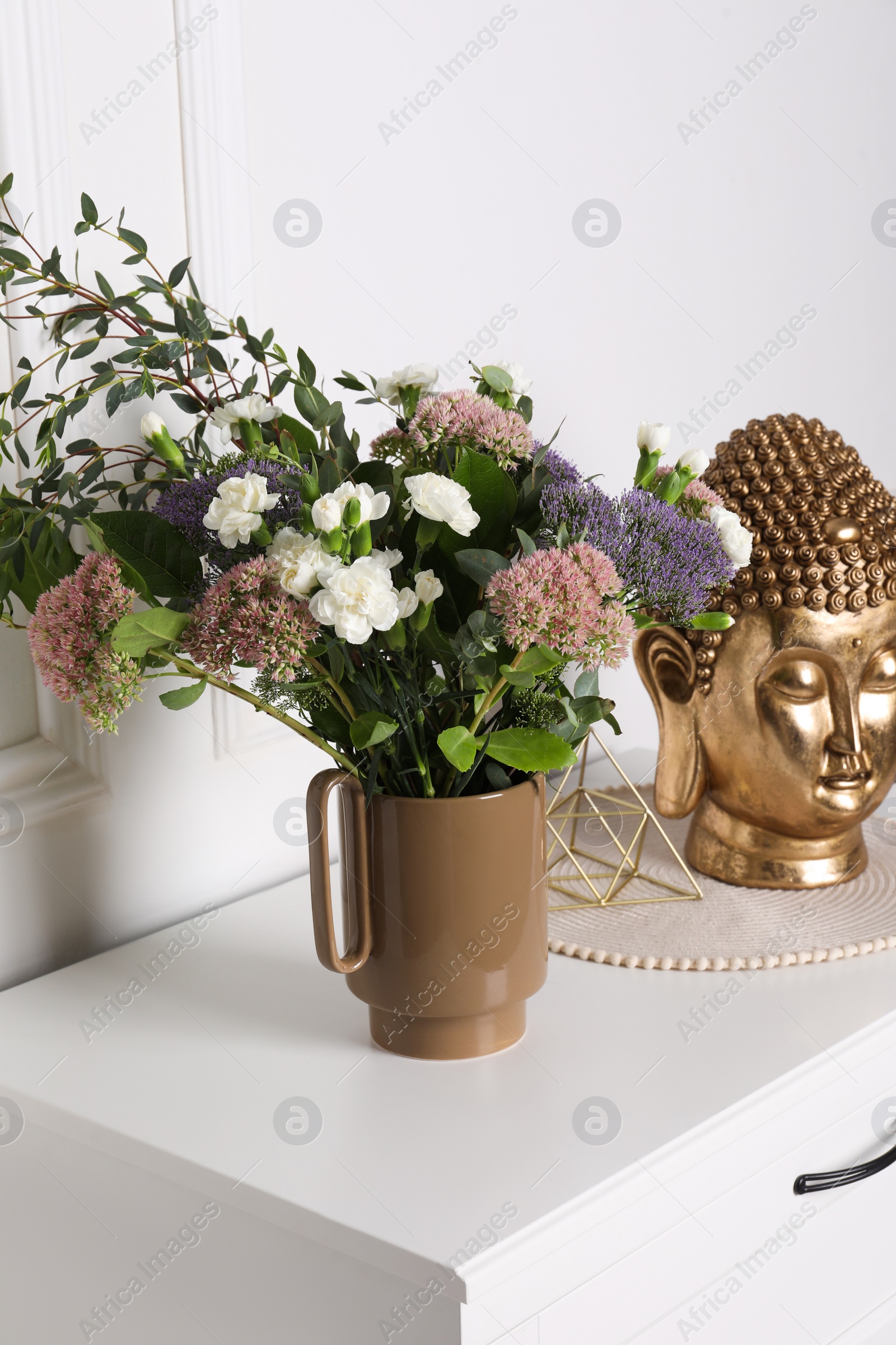 Photo of Stylish ceramic vase with beautiful flowers and golden Buddha sculpture on chest of drawers near white wall