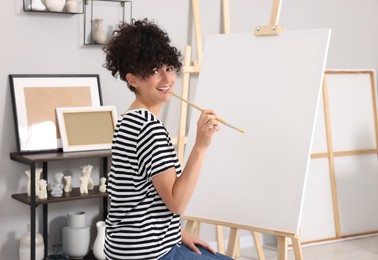 Photo of Young woman holding brush near easel with canvas in studio