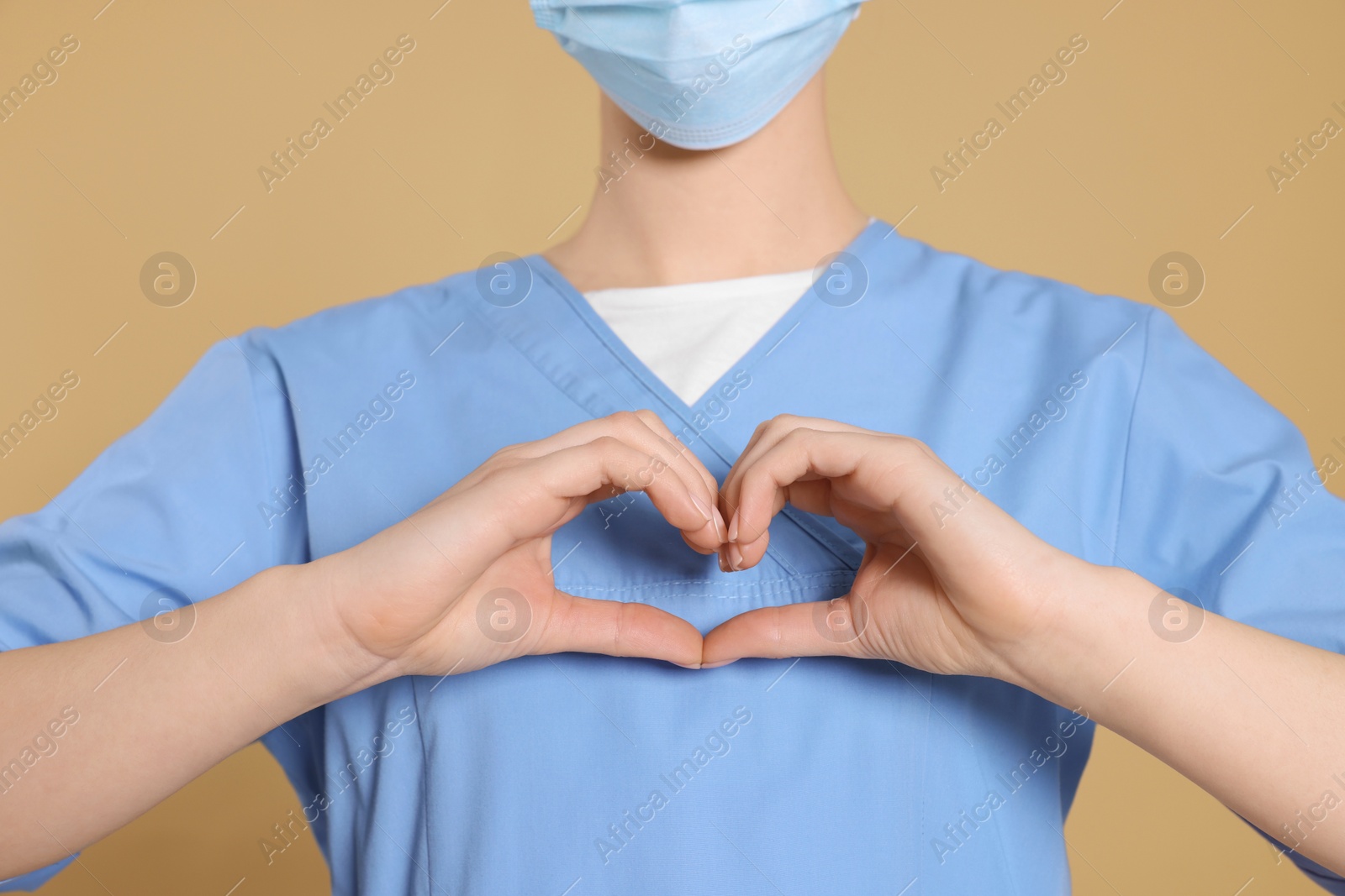 Photo of Nurse wearing protective mask and medical uniform making heart with hands on light brown background, closeup