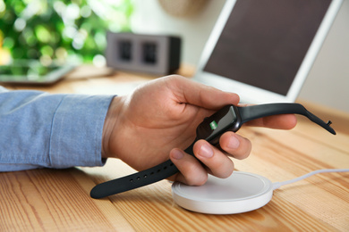 Photo of Man taking smartwatch from wireless charger at wooden table, closeup. Modern workplace accessory