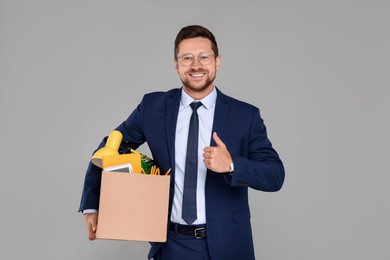 Photo of Happy unemployed man with box of personal office belongings showing thumb up on grey background