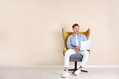 Photo of Handsome young man with laptop in armchair near color wall indoors. Space for text