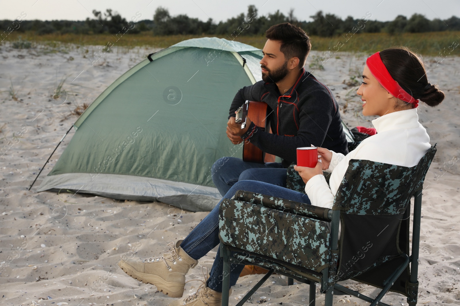 Photo of Young man playing guitar to his beloved girlfriend near camping tent on beach