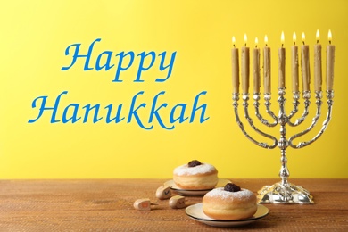 Image of Happy Hanukkah. Silver menorah, dreidels and sufganiyot on wooden table against yellow background,