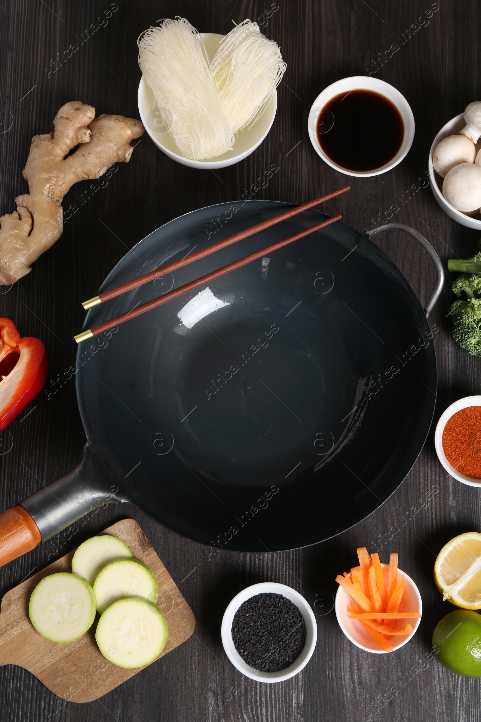 Photo of Empty iron wok and chopsticks surrounded by ingredients on dark grey wooden table, flat lay