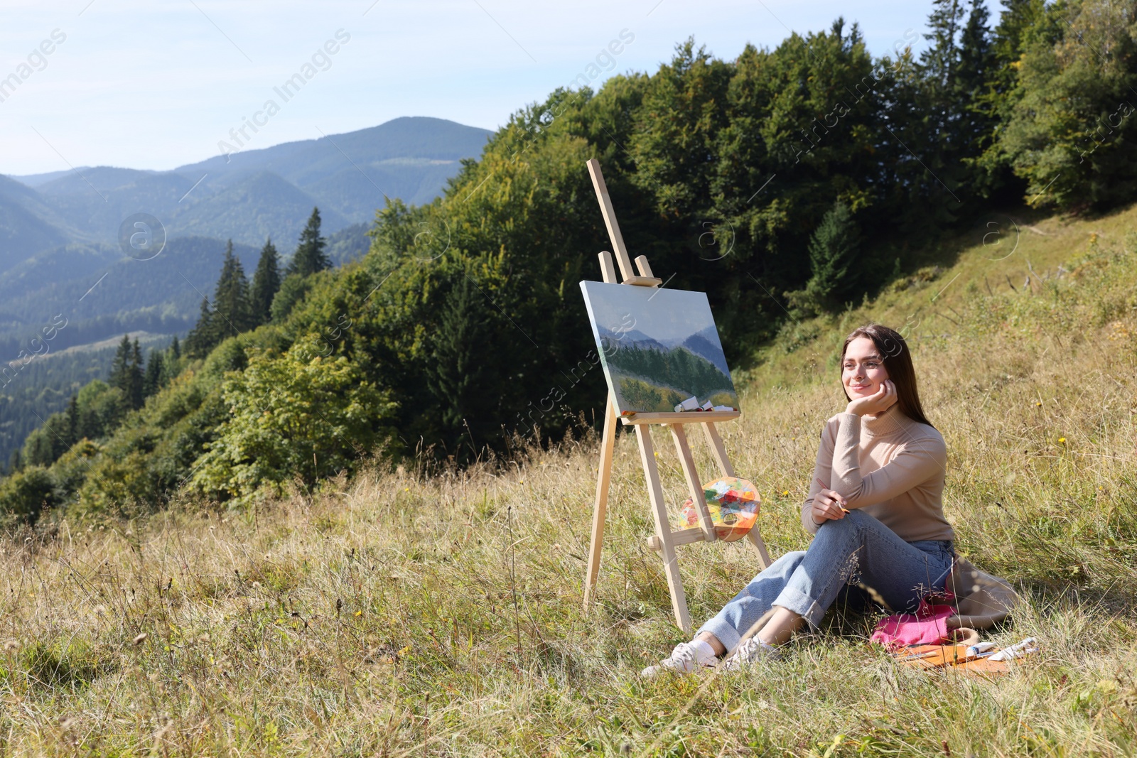 Photo of Young woman near easel with drawing in mountains, space for text