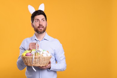 Photo of Man in cute bunny ears headband holding wicker basket with Easter eggs on orange background. Space for text
