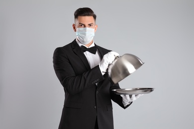 Waiter in medical face mask with empty tray on light grey background