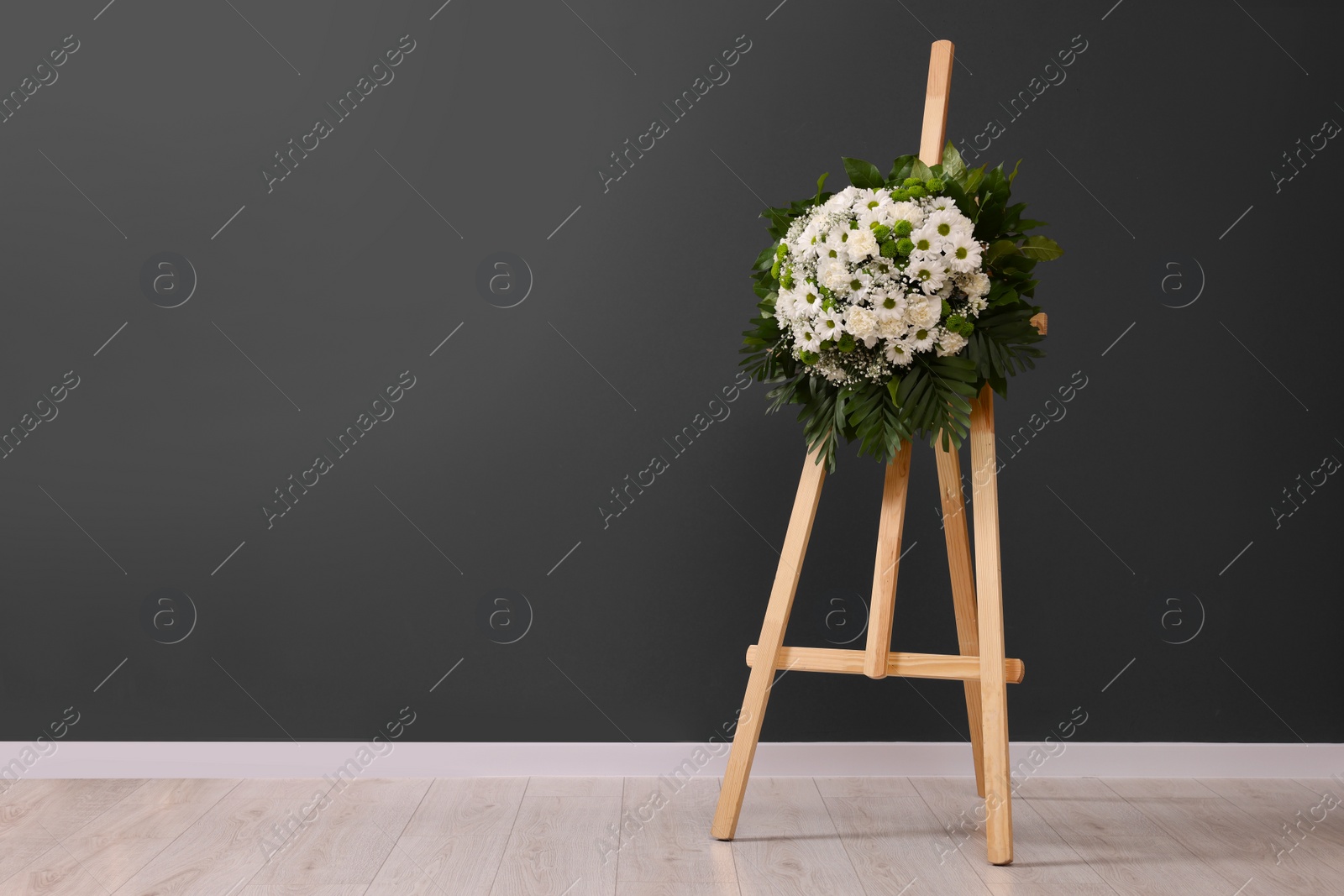 Photo of Funeral wreath of flowers on wooden stand near dark grey wall indoors, space for text