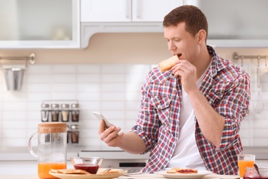 Photo of Young man eating tasty toasted bread while using mobile phone at table in kitchen