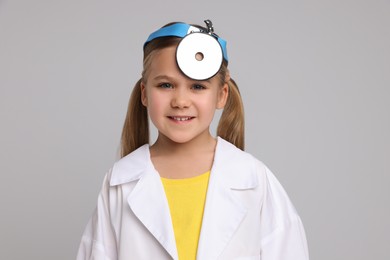 Photo of Little girl in medical uniform with head mirror on light grey background