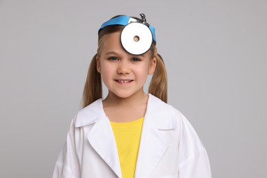 Little girl in medical uniform with head mirror on light grey background