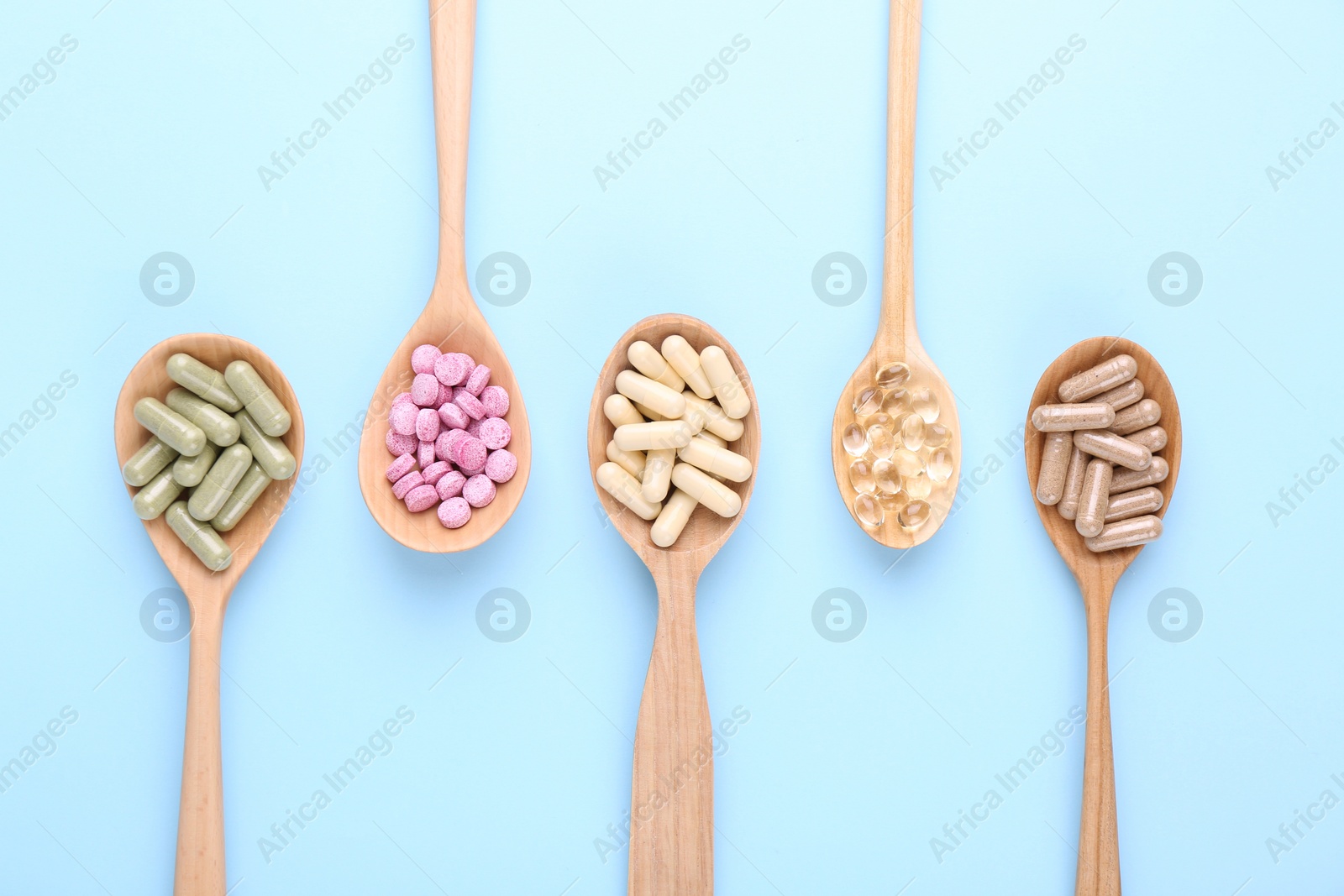 Photo of Different vitamin pills in wooden spoons on light blue background, flat lay