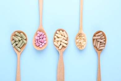 Different vitamin pills in wooden spoons on light blue background, flat lay