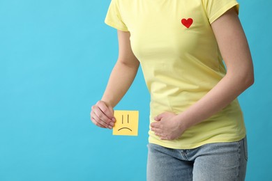 Woman holding sticky note with drawn sad face and suffering from cystitis on light blue background, closeup. Space for text