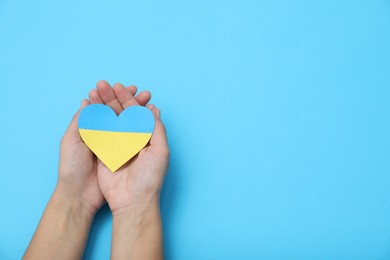 Photo of Woman holding paper heart in colors of Ukrainian flag on light blue background, top view. Space for text