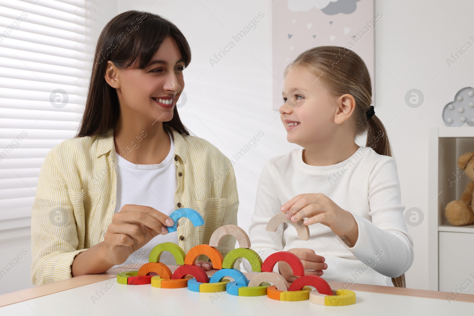 Photo of Motor skills development. Mother helping her daughter to play with colorful wooden arcs at white table in room