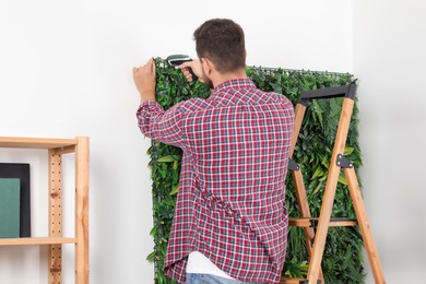 Man with screwdriver installing green artificial plant panel on white wall in room, back view