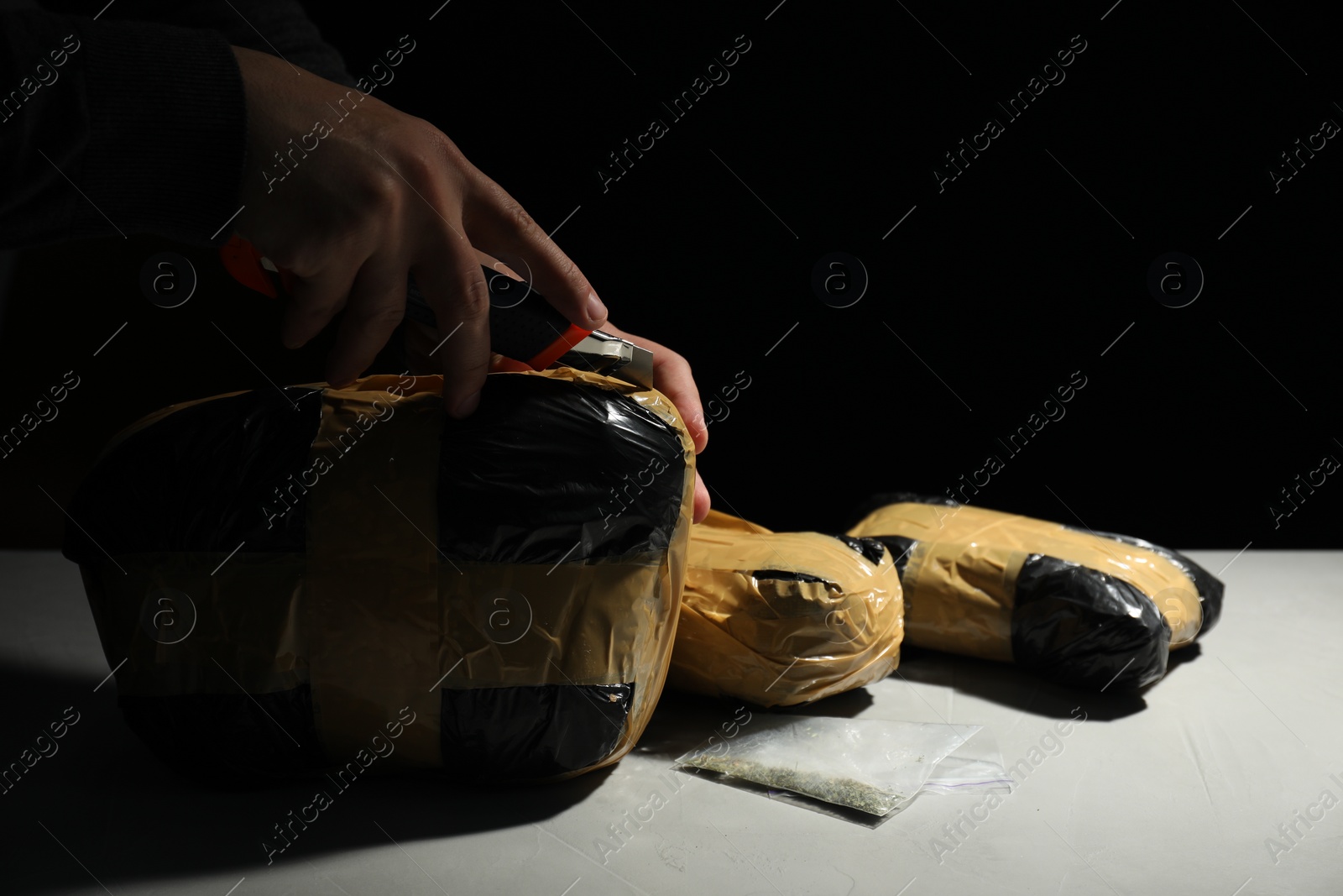 Photo of Smuggling and drug trafficking. Man opening package of narcotics with box cutter at light table against black background, closeup