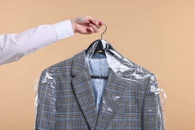 Photo of Dry-cleaning service. Woman holding jacket in plastic bag on beige background, closeup